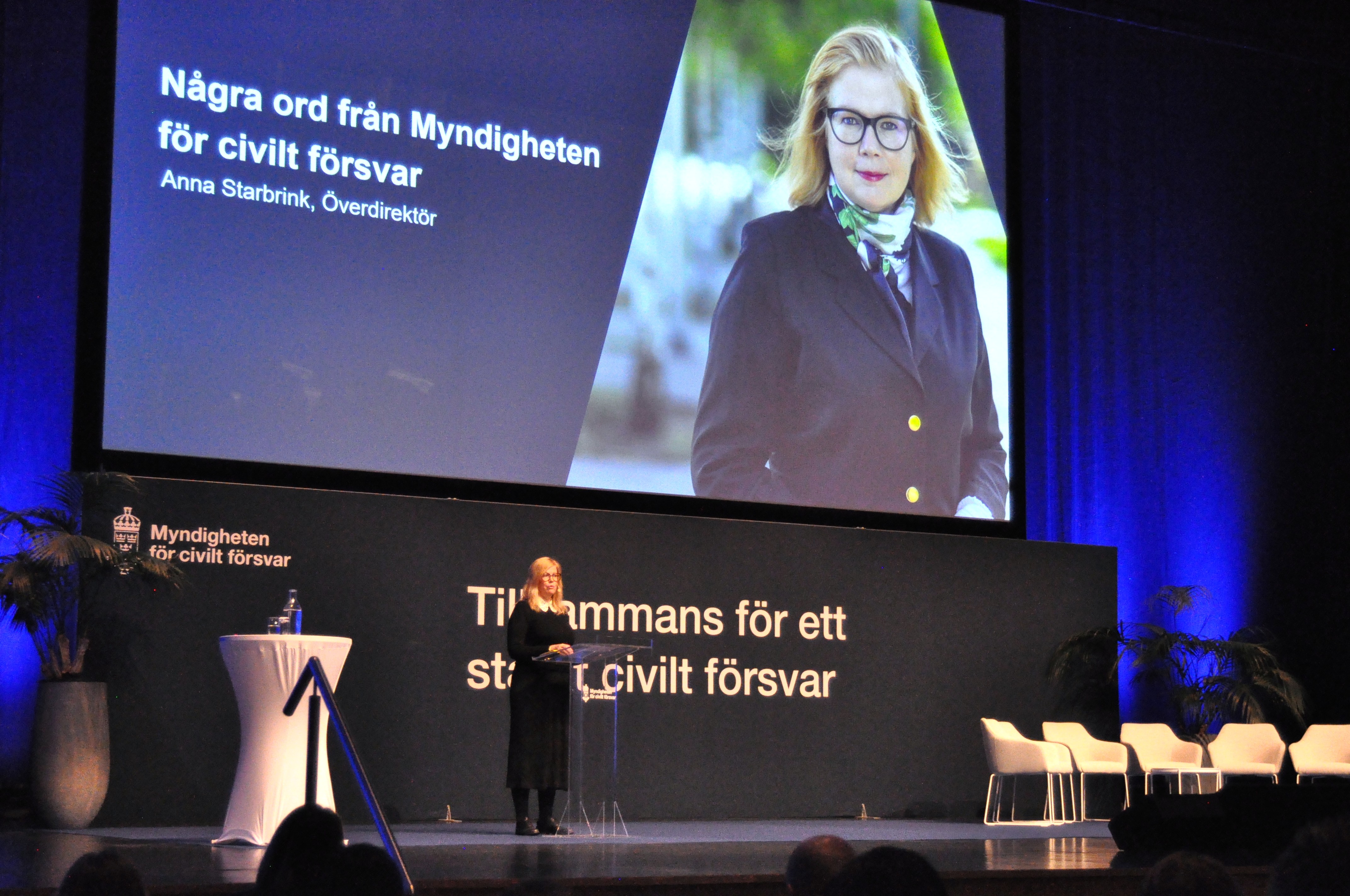 Överdirektör på Myndigheten för civilt försvar Anna Starbrink står på en scen och talar till publiken. Bak henne finns en stor skärm med en bild på överdirektören och texten Några ord från Myndigheten för civilt försvar.